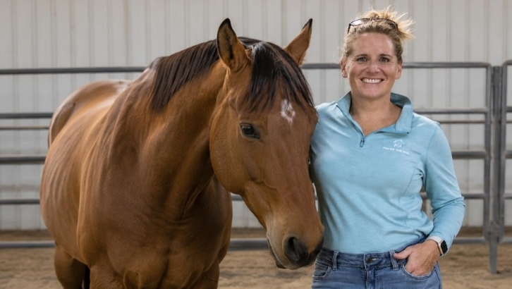 Woman standing beside a brown horse inside an equine facility.